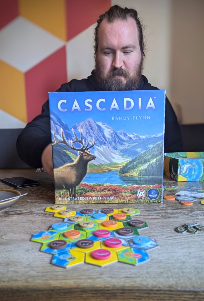 A photo of the Cascadia box and game set up on the table at the board game cafe. Matt is sat behind the box looking more serious than normal! (Probably reading the rules!)